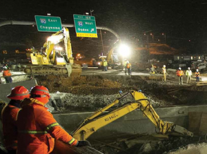 Night photo of a Flatiron construction crew moving a bridge. Two crew members are shown wearing orange jackets. There are tractors in the background.